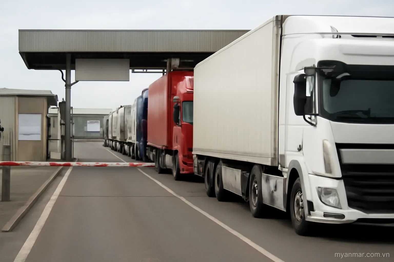Trucks queued at a border checkpoint with barriers and warehouses, wide view, no text